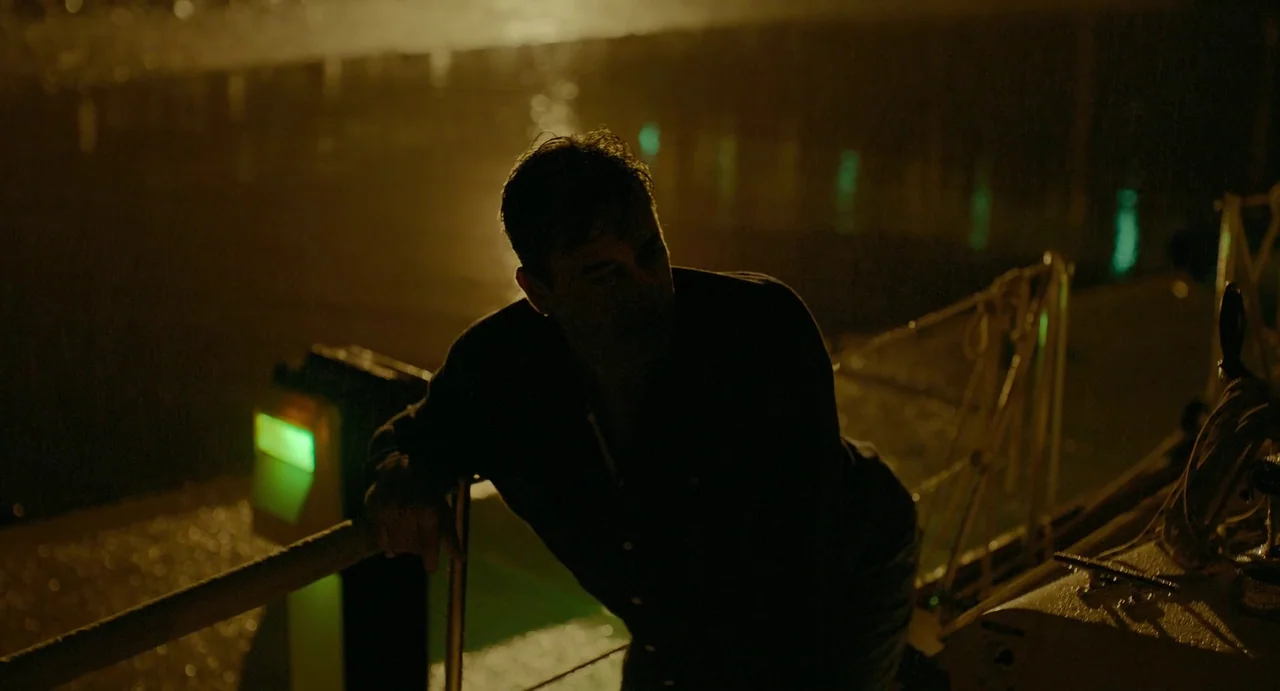 Confess, Fletch (2022) movie still: medium — A man stands on the deck of a boat at night near dark water. He leans against a metal railing as green…