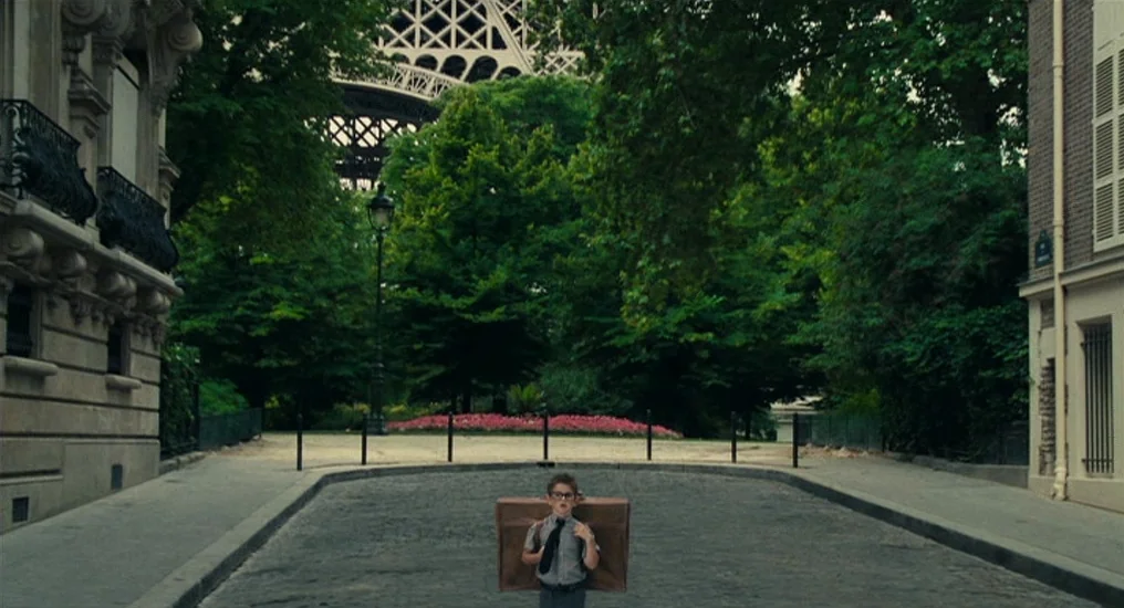 Paris, Je T’Aime (2006) movie still: wide — A quiet cobblestone street in Paris features the Eiffel Tower visible in the background. A small boy…