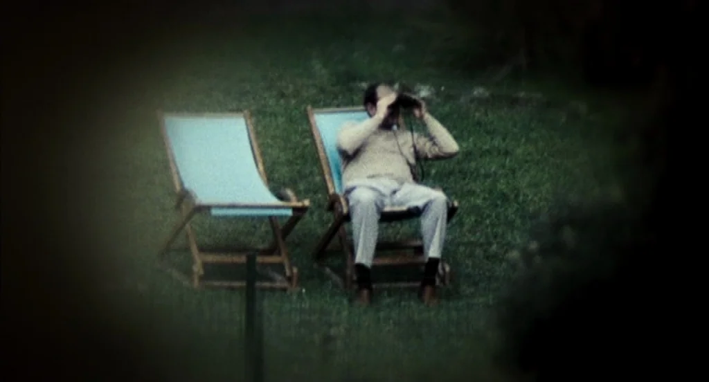 Timecrimes (2007) movie still: wide — A man sits on a lawn in a wooden beach chair with binoculars. An empty matching chair is placed next to him on…
