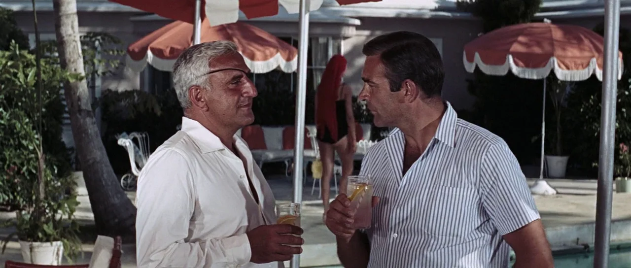 Thunderball (1965) movie still: medium — A sunny poolside patio features large red and white umbrellas shading the area. Two men in collared shirts…