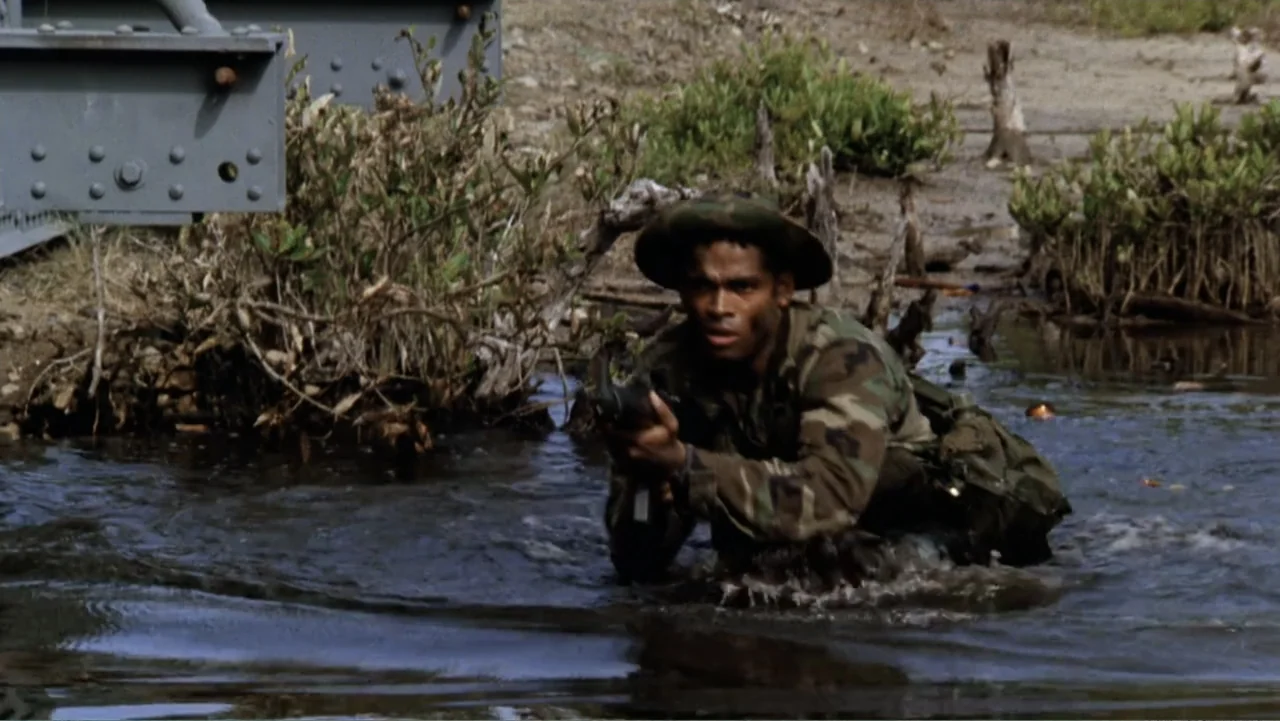 Heartbreak Ridge (1986) movie still: medium — A soldier in military camouflage wades through a murky swamp under an iron structure. The individual…