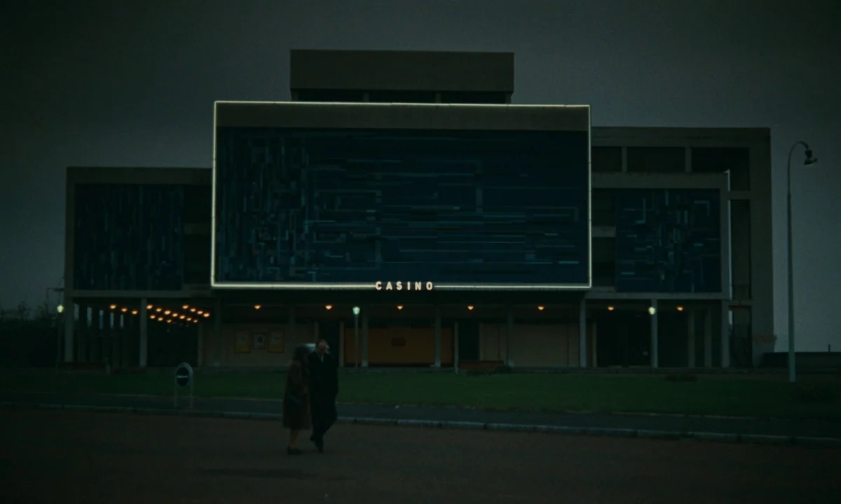 Muriel (1963) movie still: establishing — A brutalist concrete casino building features a prominent geometric mural and a neon sign. Two figures…