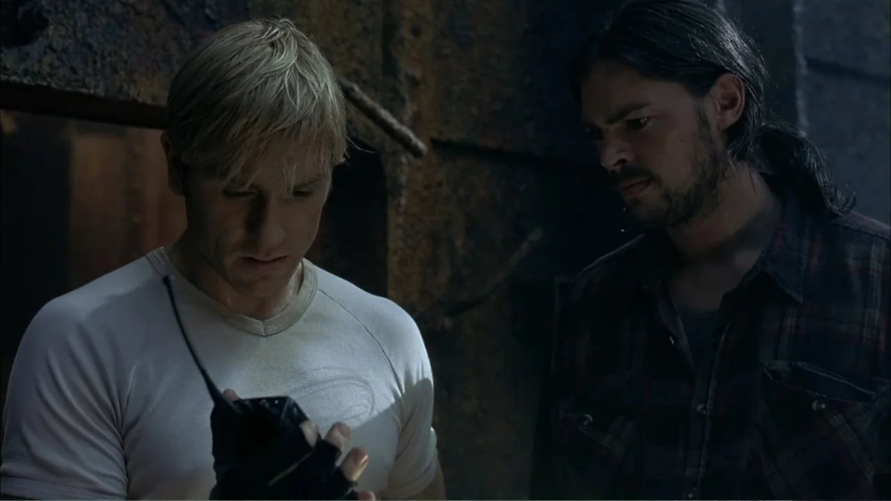 Ghost Ship (2002) movie still: medium — A dark, dimly lit industrial space with textured metal walls and rusted surfaces. Two men in casual clothing…