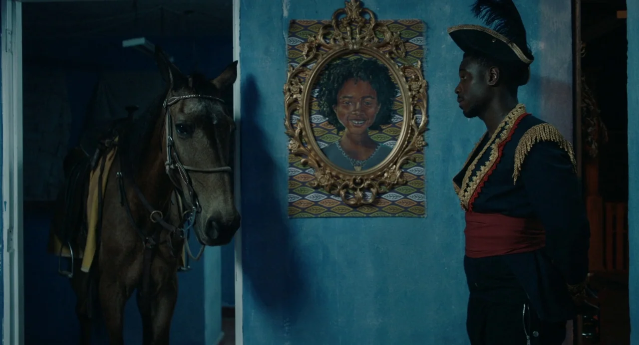 The Day Shall Come (2019) movie still: medium — A blue hallway features a framed portrait of a girl between two doorways. A saddled horse stands on…