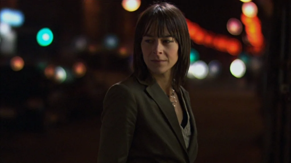 Red Road (2006) movie still: medium — A woman stands on a city street at night with out-of-focus city lights behind her. She looks over her shoulder…