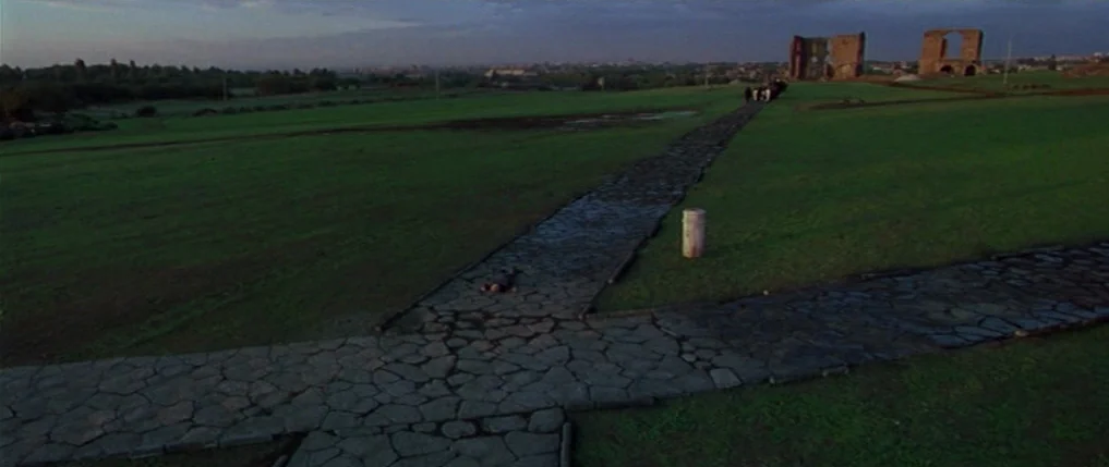 Titus (1999) movie still: establishing — A wide shot shows an expansive green park with ancient masonry structures in distance. A small person is…