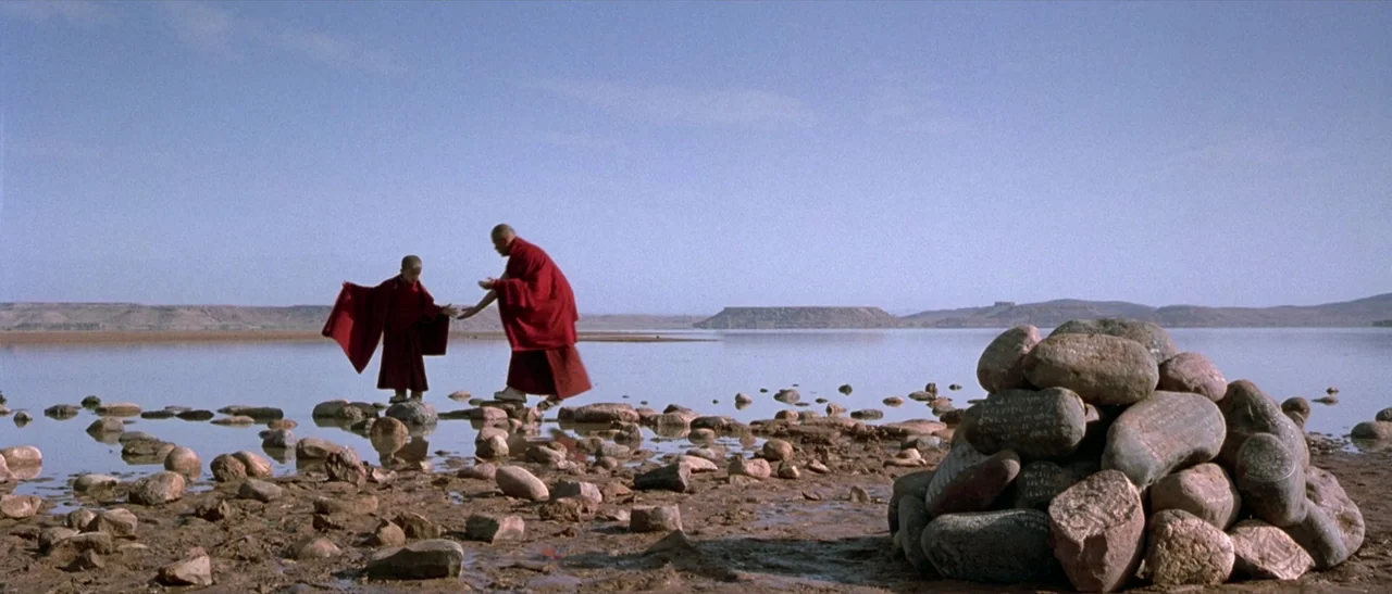 Kundun (1997) movie still: wide — A vast, shallow lake stretches toward distant hills under a clear blue sky. Two Buddhist monks in vibrant red…