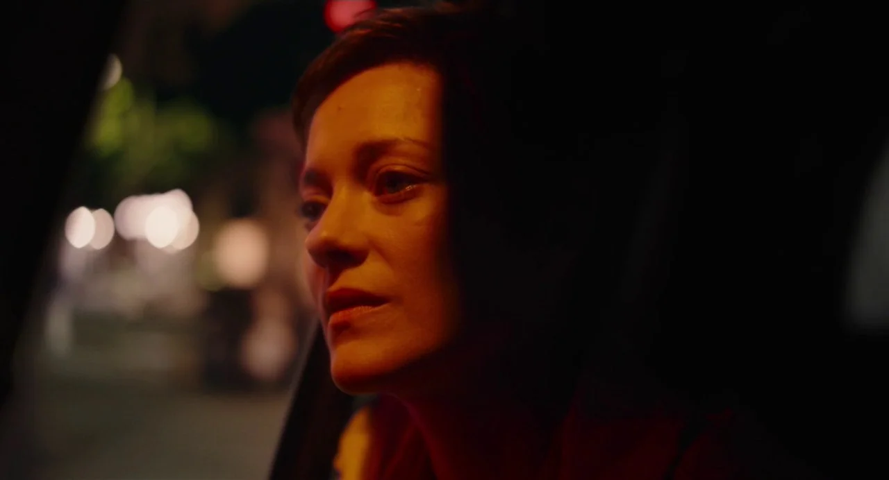 Annette (2021) movie still: closeup — The interior of a vehicle at night featuring a woman looking out a window. Outside the window, out-of-focus…