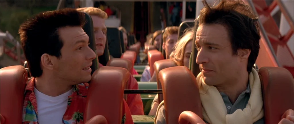 True Romance (1993) movie still: medium — A roller coaster car travels along tracks in an amusement park setting. Two men in the front row face each…