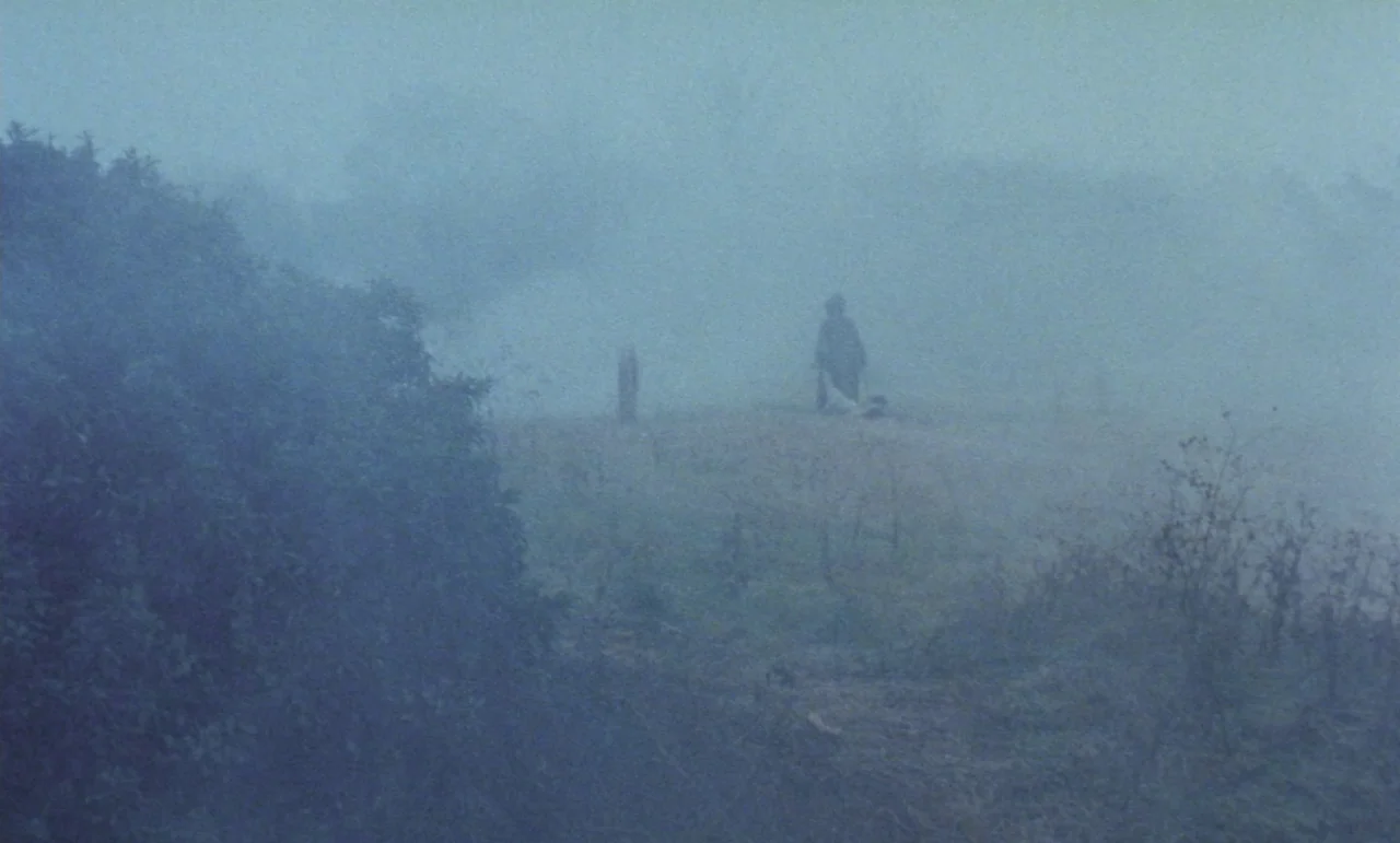 Evil Dead Trap (1988) movie still: wide — A desolate field covered in a dense, heavy layer of morning fog. A single dark, hooded figure stands…