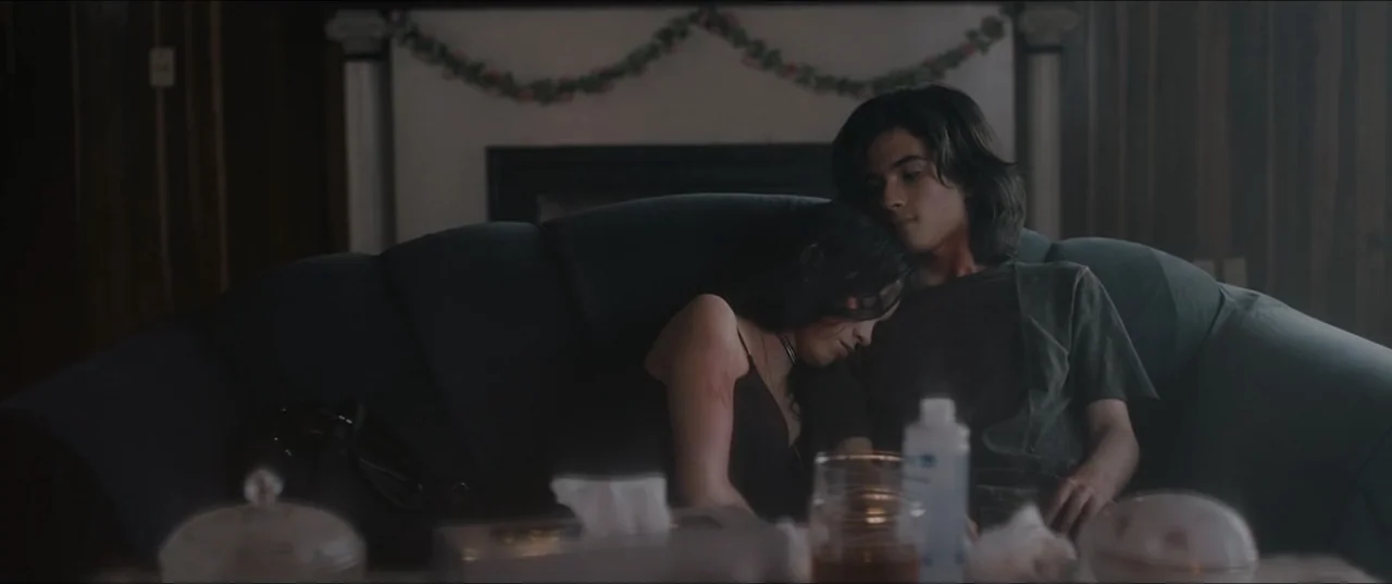 Death to Summer (2021) movie still: medium — A dimly lit room with a dark couch featuring a decorative garland on the wall. A young man and woman…