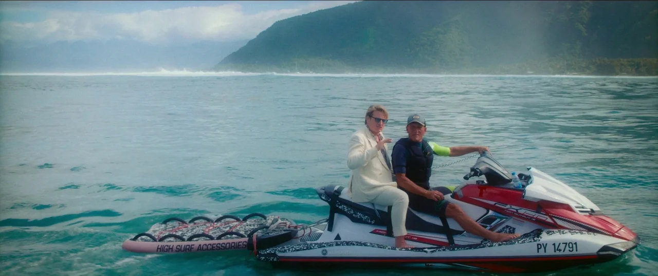 Pacifiction (2022) movie still: wide — A calm tropical ocean with mountains in the background under an overcast sky. Two men sit on a red and white…