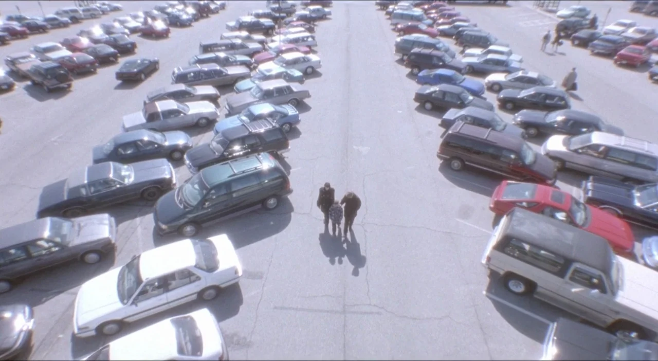 Twelve Monkeys (1995) movie still: wide — A vast, asphalt parking lot filled with various 1990s-era automobiles arranged in orderly rows. Three…