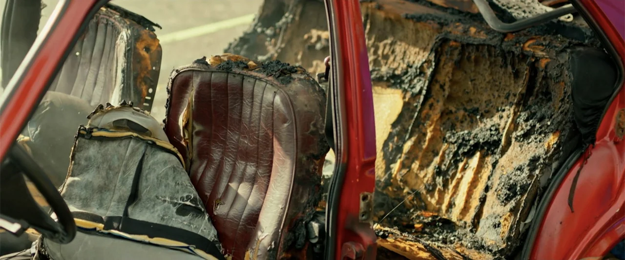 Wrong Package (2025) movie still: medium — The interior of a fire-damaged vehicle is exposed through an open door. Scorched upholstery and charred…