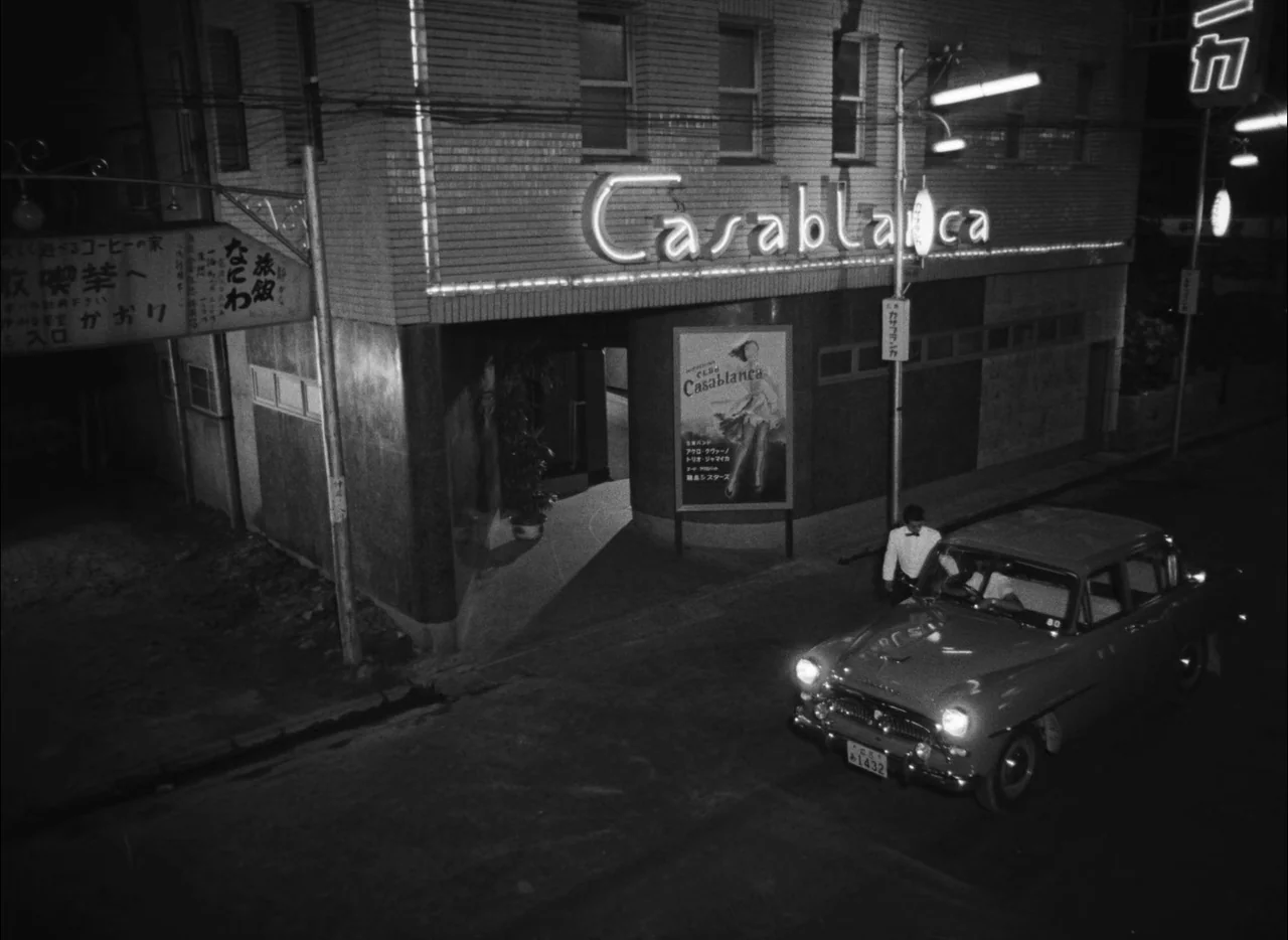 Hiroshima Mon Amour (1959) movie still: wide — A dark night street corner in Japan features a building titled Casablanca. A vintage sedan drives…