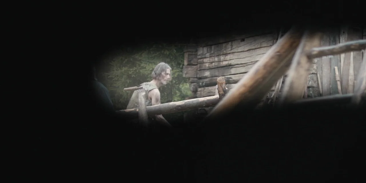 The Ritual (2017) movie still: medium — A man stands outdoors near the logs of a weathered wooden cabin. He holds a large manual crosscut saw while…