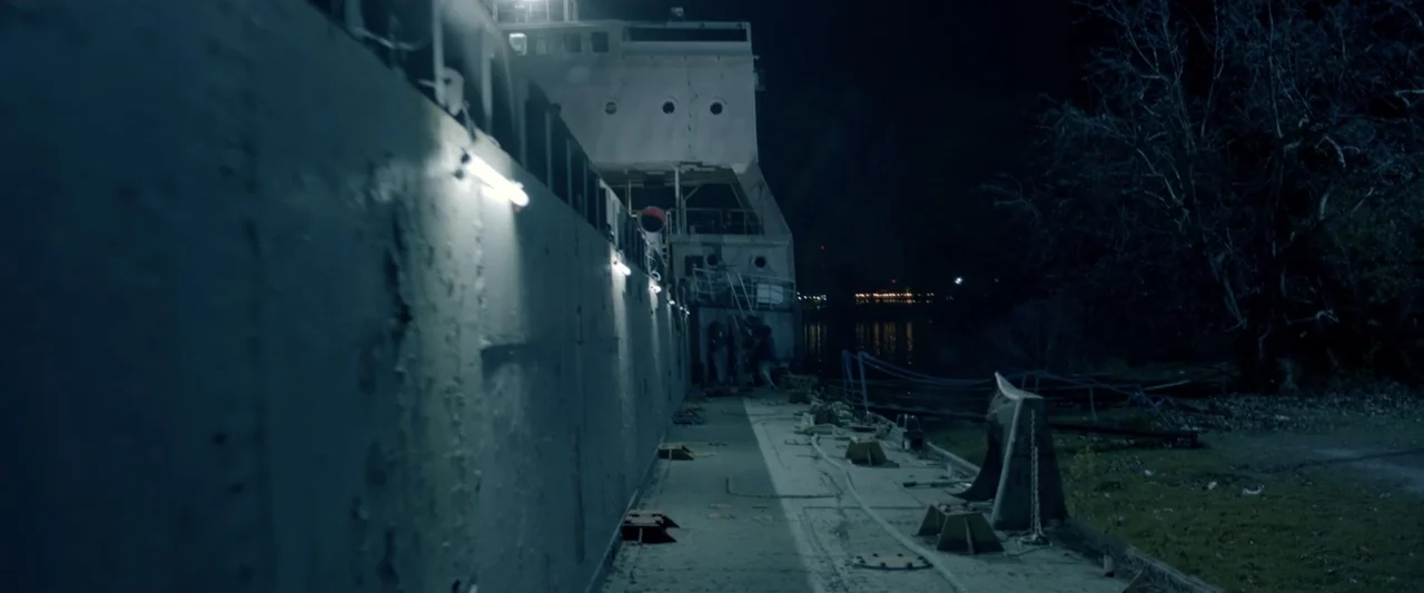 Perdida (2018) movie still: wide — A dark, deserted ship deck at night is illuminated by a series of fluorescent lights. The metal hull of the…