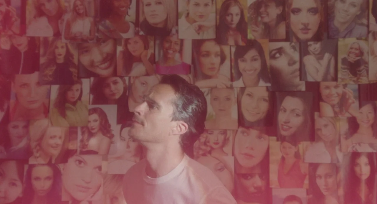 Babysitter (2022) movie still: medium — A wall is densely covered with numerous printed photographs of various women's faces. A man stands in the…