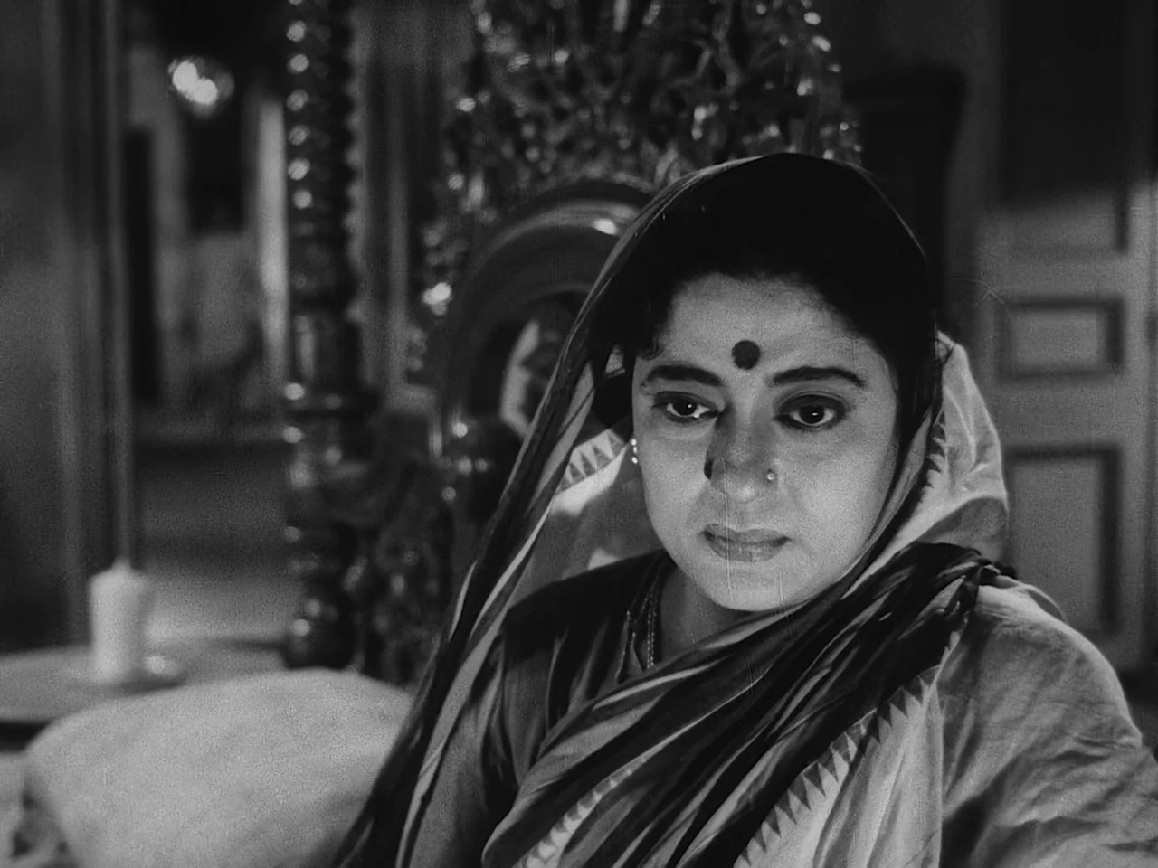 The Music Room (1958) movie still: closeup — A woman sits in a room with a traditional carved wooden bed headboard behind. She looks away from the…