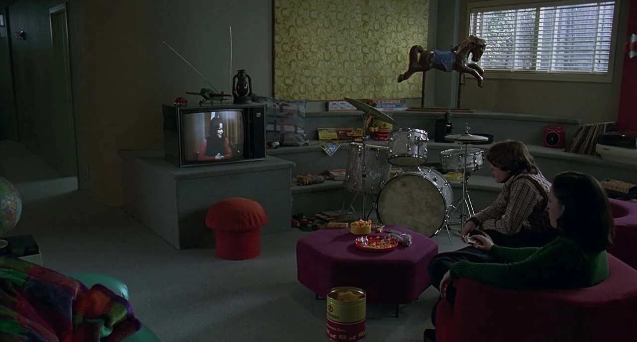 The Ice Storm (1997) movie still: wide — A basement room with textured walls and a raised tier featuring a drum kit. A teenage boy and girl sit on…