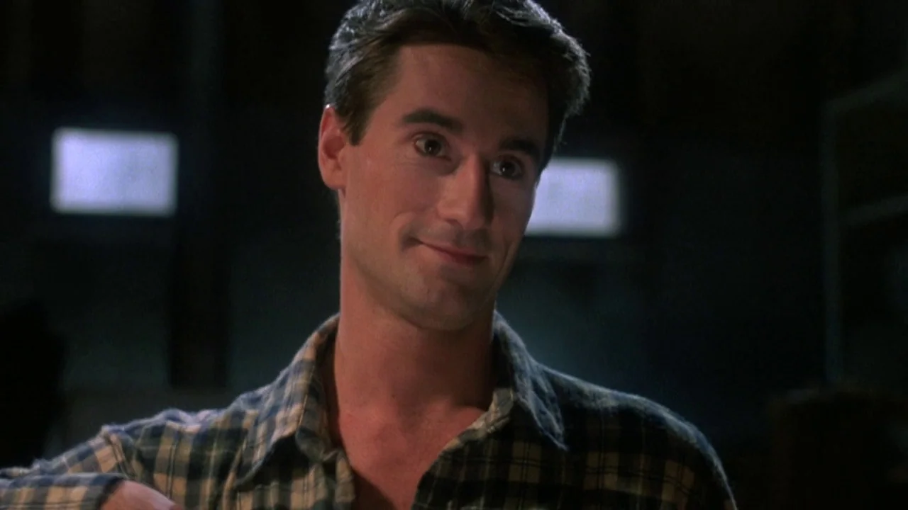 Re-Animator (1985) movie still: medium — A man in a dark plaid shirt stands in a dimly lit, undefined indoor space. He is looking off-camera while…