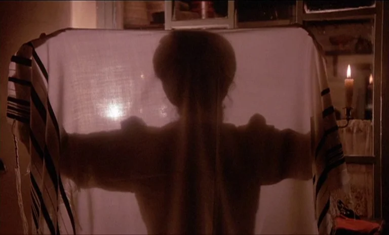 Yentl (1983) movie still: medium — A dark indoor setting features a person holding up a patterned white cloth. The backlit figure is seen through…