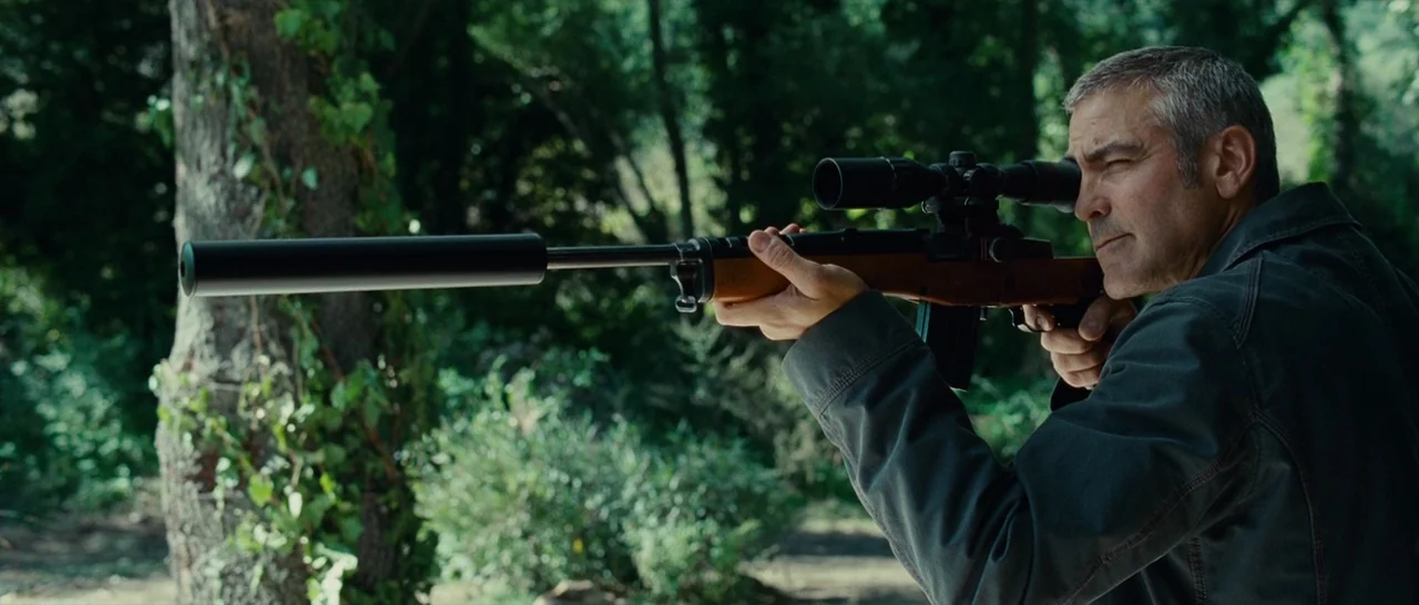 The American (2010) movie still: medium — A man stands in a dense wooded area with lush green foliage behind him. He holds a black suppressed rifle…