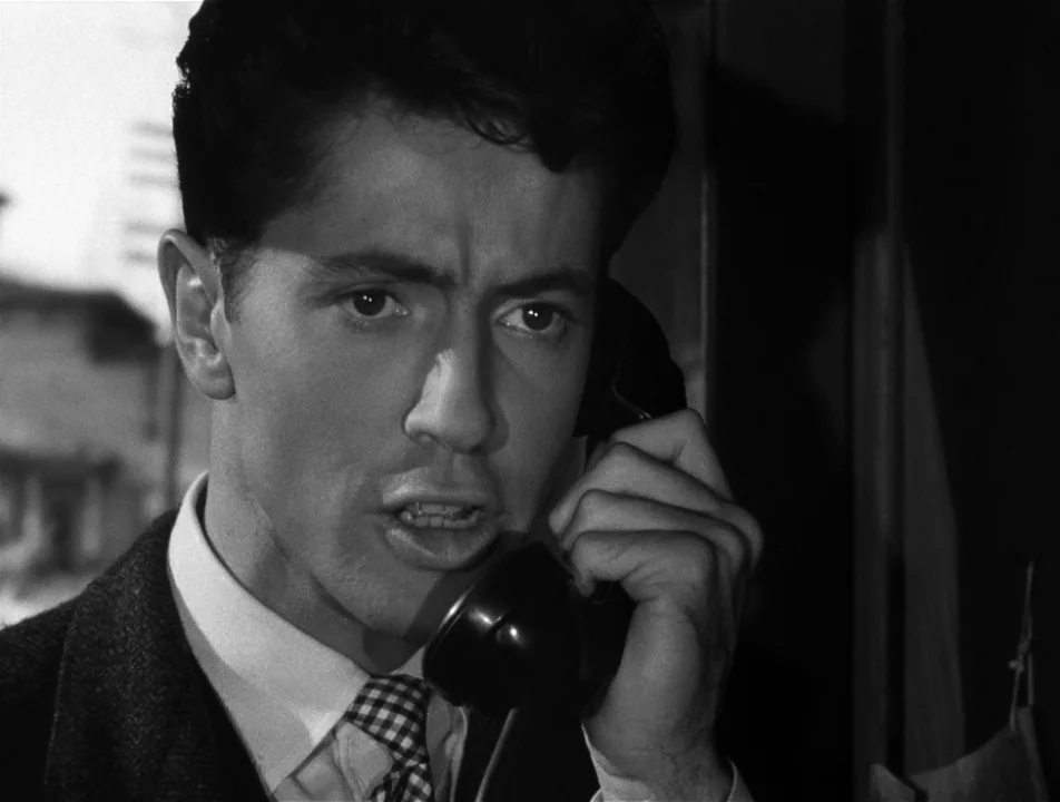 Strangers on a Train (1951) movie still: closeup — A man speaks on a telephone inside a small, cramped enclosure. He looks forward with a furrowed…