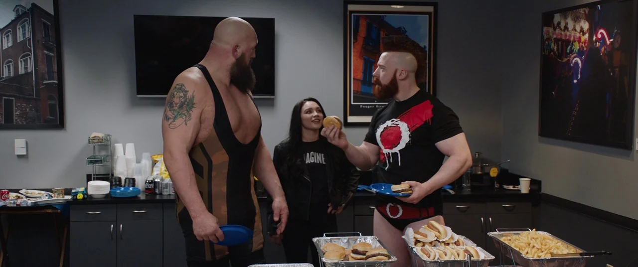 Fighting With My Family (2019) movie still: medium — A break room features black cabinets and catering trays filled with fast food items. Two…
