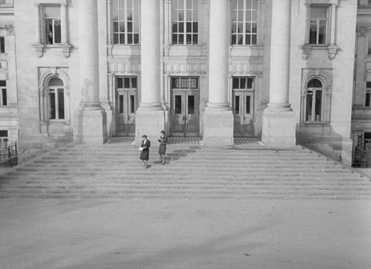 Wings (1966) movie still: wide — A grand neoclassical building features tall columns and symmetrical glass-paneled wooden doors. Two women walk…