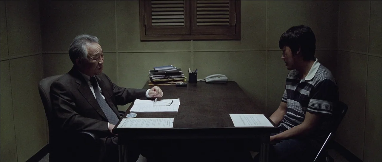 The Chaser (2008) movie still: medium — An austere room features plain green paneled walls and a shuttered wooden window. Two men face each other…