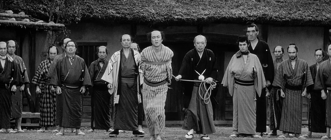 Yojimbo (1961) movie still: fullbody — A group of Japanese men gather in front of a rustic wooden building with thatch. A man bound in rope stands…