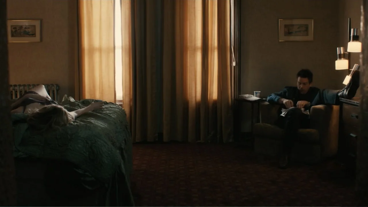Maggie's Plan (2016) movie still: wide — A dimly lit hotel room features a bed covered in a green quilt and furniture. A woman rests on the bed…