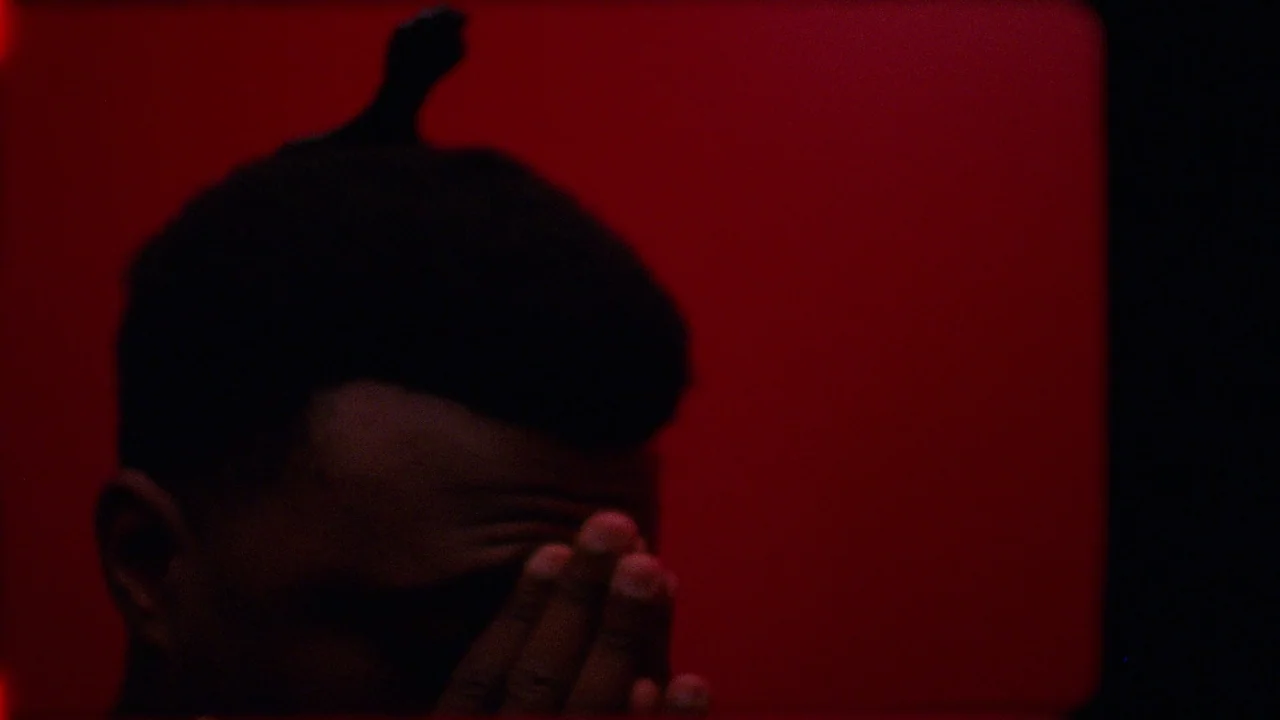 Pass Over (2018) movie still: closeup — A person stands in a dimly lit space against a flat red background. The subject holds their hands over their…