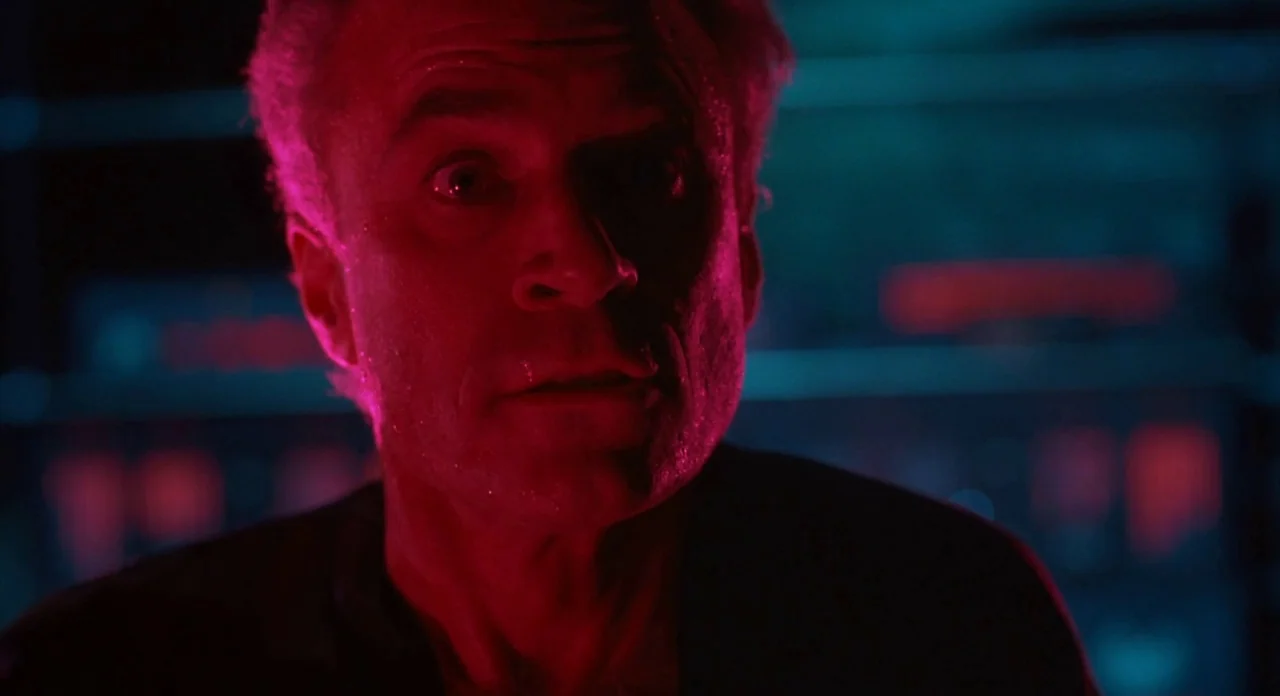 From Beyond (1986) movie still: closeup — A man's face is illuminated in a dark, neon-lit interior space. He stares intensely toward the camera…