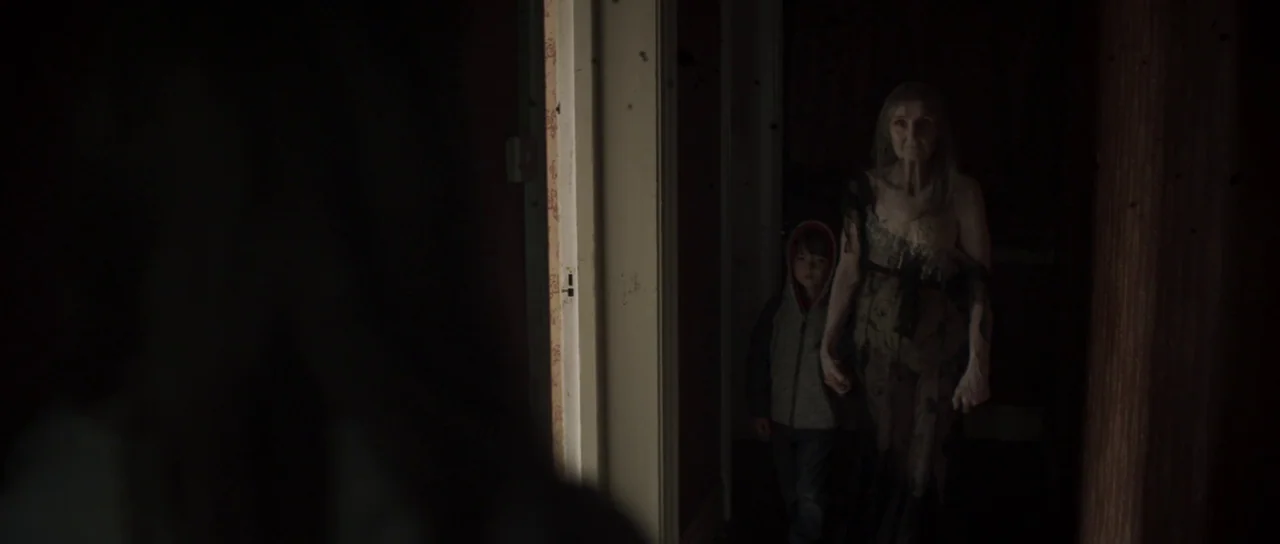 A Dark Song (2016) movie still: medium — A dark, narrow residential corridor with plain walls and a slightly open door frame. An old woman and a…