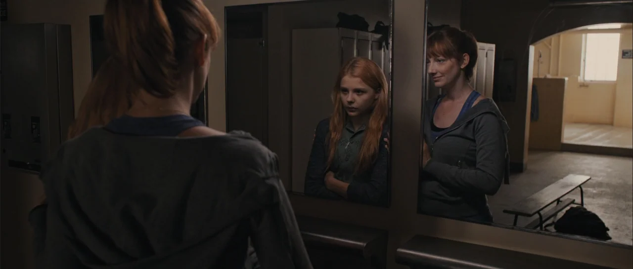 Carrie (2013) movie still: medium — A utilitarian locker room features metal lockers and a long horizontal wall mirror. An older woman stands behind…
