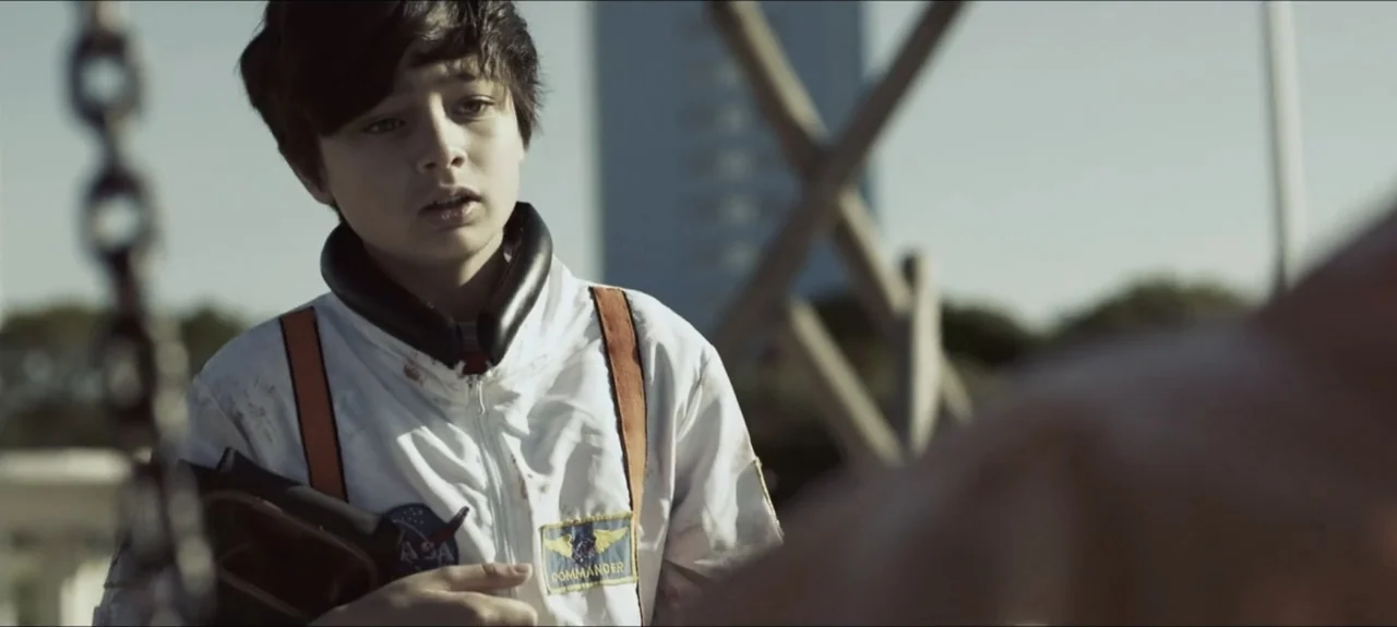 Containment (2015) movie still: medium — A playground with blurred wooden structures and a tall building in the distance. A young boy wearing an…