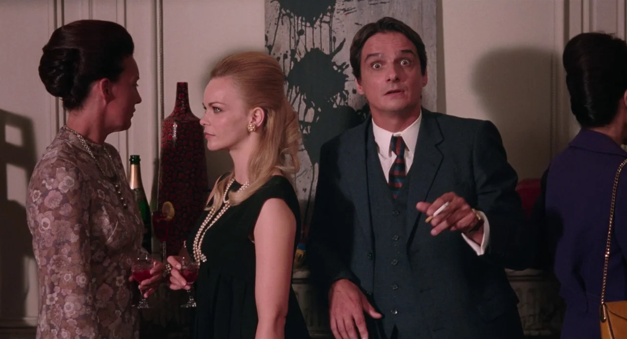Godard Mon Amour (2015) movie still: medium — A well-appointed 1960s parlor with paneled walls and an abstract painting hanging behind the group. A…