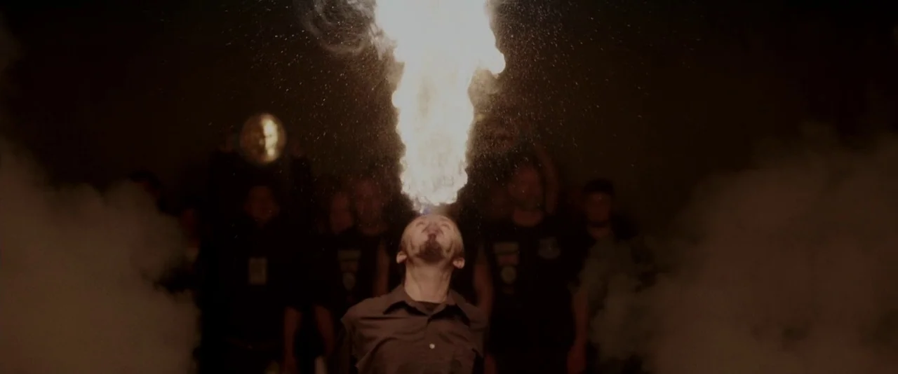 Creed (2015) movie still: medium — A man stands in a dark, hazy outdoor crowd while performing a fire-breathing act. A thick column of orange flames…