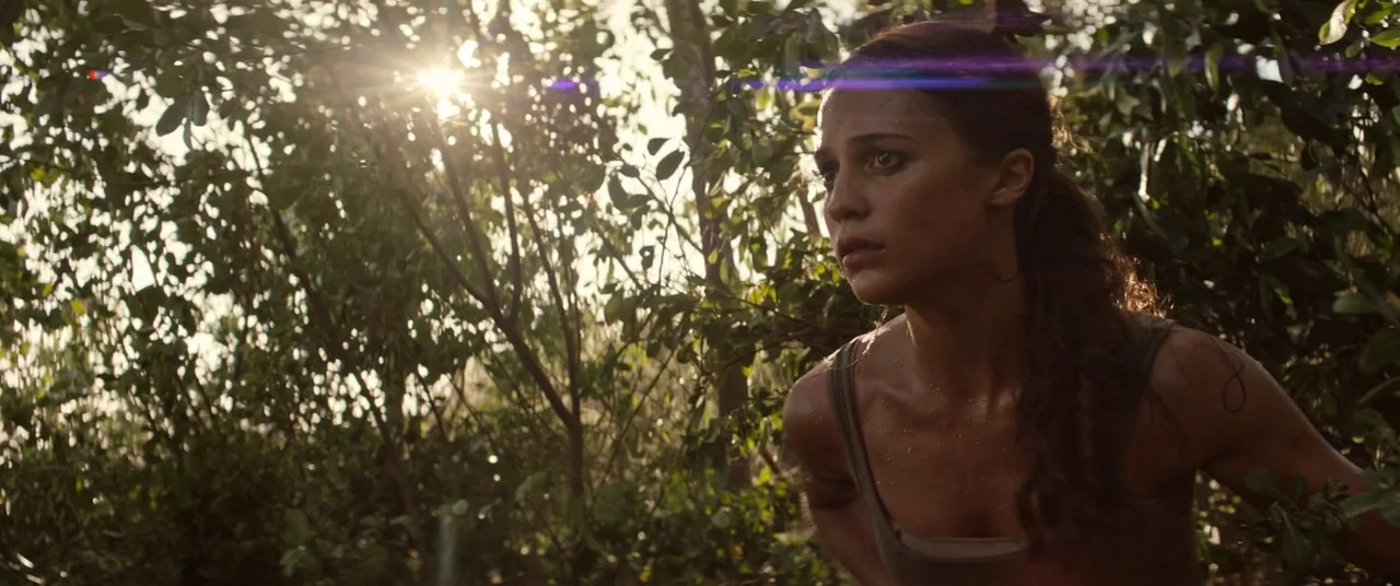 Tomb Raider (2018) movie still: medium — Dense tropical vegetation frames the shot with bright sunlight filtering through the leaves above. A woman…