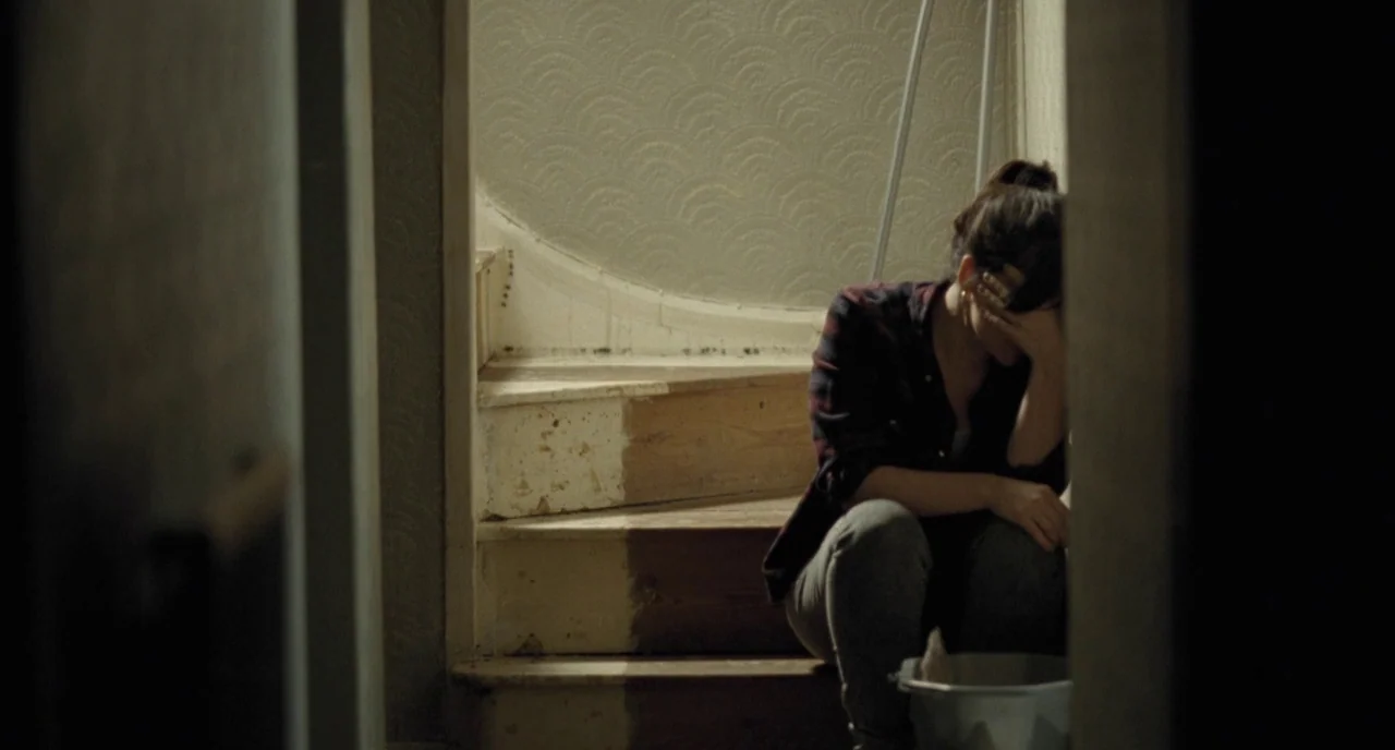 I, Daniel Blake (2016) movie still: medium — A dimly lit staircase features worn wooden steps and wallpaper with a subtle pattern. A woman sits on…