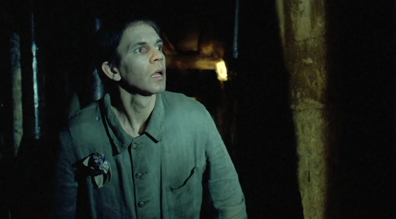 Underground (1995) movie still: medium — A dark subterranean cavern with rugged rock walls visible behind the subject. A man stands in the low light…