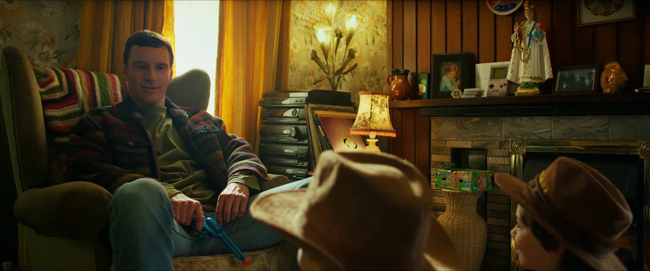 Kneecap (2024) movie still: medium — A cluttered 1990s living room features wood-paneled walls and a tiled fireplace mantle. An adult man sits in an…