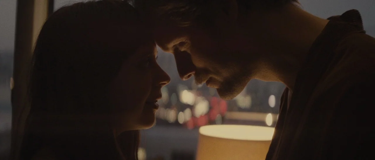 The Diary of a Teenage Girl (2015) movie still: closeup — A dimly lit room overlooks a blurred city skyline at night through a window. A man and a…