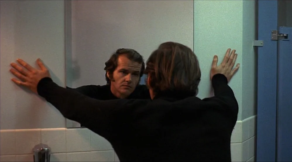 Five Easy Pieces (1970) movie still: medium — A utilitarian public restroom features white wall tiles and blue painted partitions. A man stands…