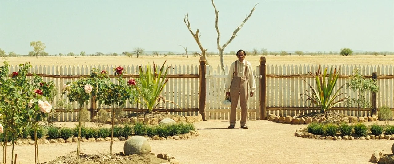 The Proposition (2005) movie still: fullbody — A man stands in an arid garden before a white picket fence in flat light. The background reveals a…