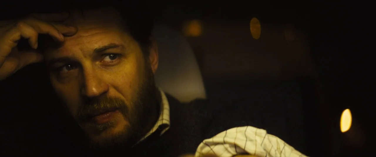 Locke (2013) movie still: closeup — Inside a dark vehicle at night with blurred city lights in the background. A man sits in the driver seat with…