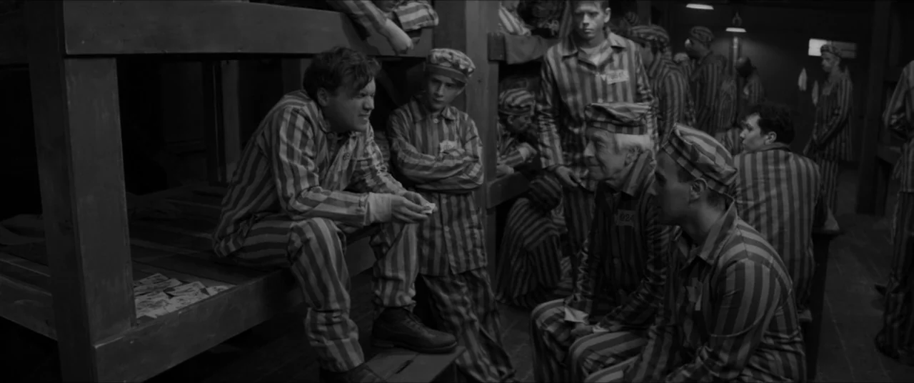 Bau, Artist at War (2025) movie still: medium — A dimly lit room contains multiple stacked wooden bunks filled with men. Several prisoners wearing…