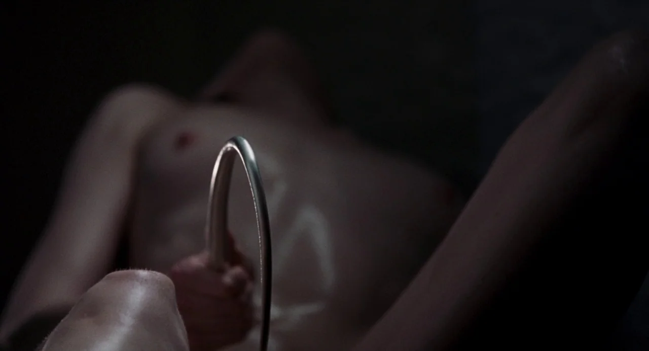 Suspiria (2018) movie still: closeup — A dark medical space is illuminated by a narrow beam highlighting a metal tool. A hand grips the curved…